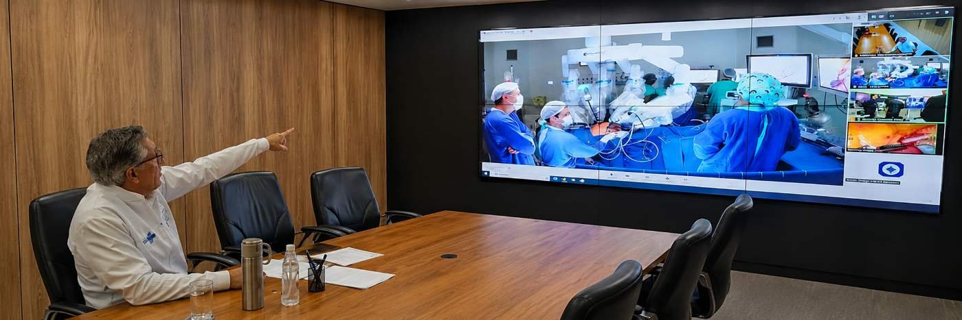 Médico acompanha em sala de conferência uma cirurgia robótica exibida em telão, representando telecirurgia entre Porto Velho e Barretos.