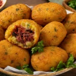 Bolinho de mandioca com carne-seca rende 50 unidades Bolinhos de mandioca dourados recheados com carne-seca, com uma unidade aberta mostrando o recheio