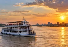 Porto Velho terá 1.500 ingressos grátis para passeios no Rio Madeira Composição editorial de barco de passageiros navegando em rio ao pôr do sol, para ilustrar passeio de barco no Rio Madeira.