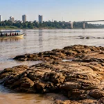 Defesa Civil alerta para riscos no Rio Madeira mesmo após chuvas em Porto Velho Composição editorial com embarcação no rio, pedras e bancos expostos que ilustram riscos à navegação no Rio Madeira.