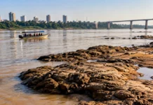 Defesa Civil alerta para riscos no Rio Madeira mesmo após chuvas em Porto Velho Composição editorial com embarcação no rio, pedras e bancos expostos que ilustram riscos à navegação no Rio Madeira.